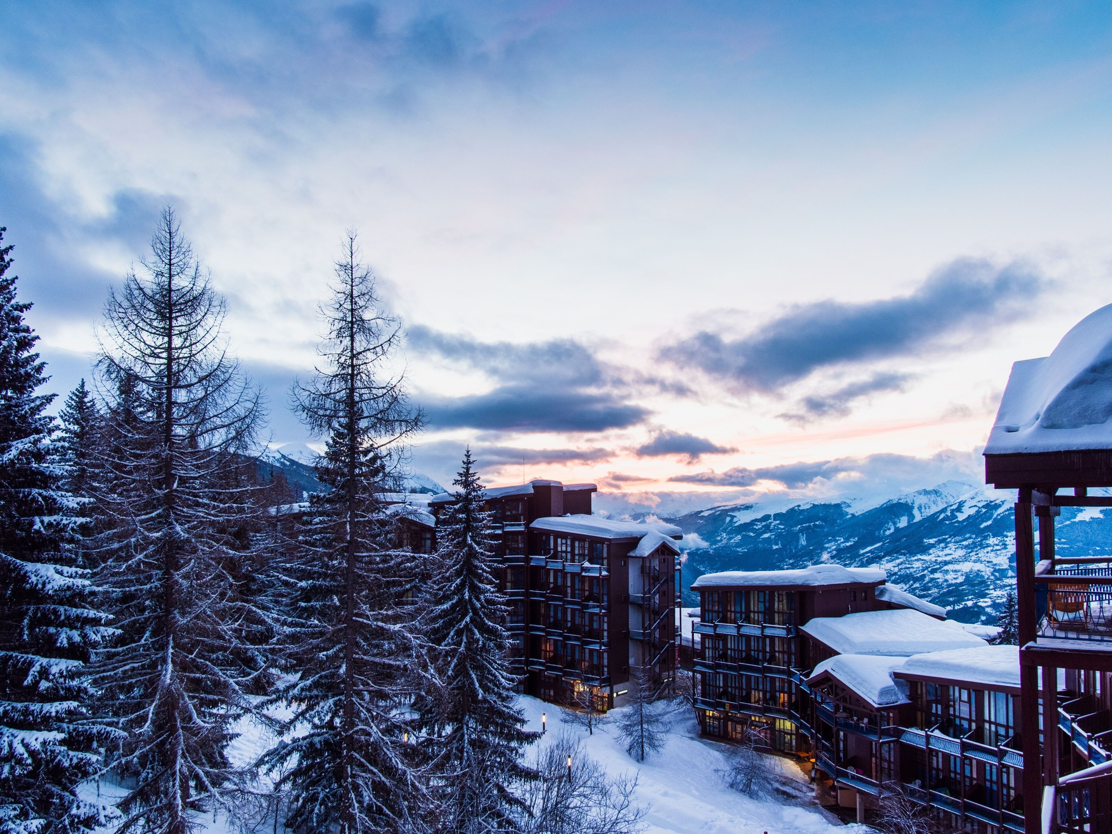 A snow-covered lodge with multiple buildings, surrounded by snow-laden trees and mountains in the background.