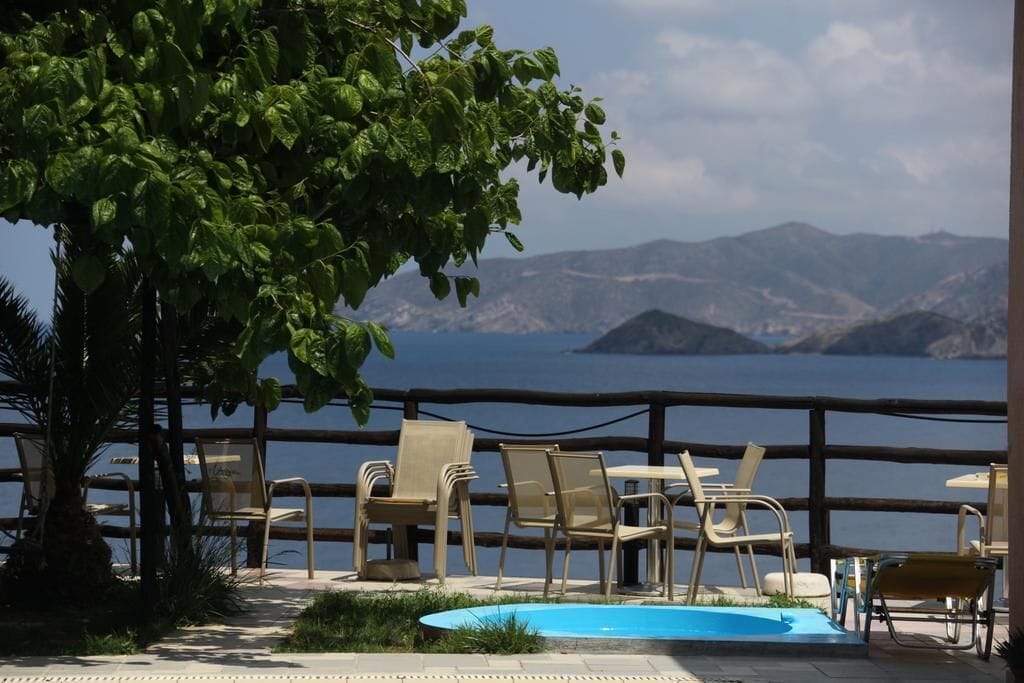 A poolside area with chairs and a table, overlooking a body of water and mountains.