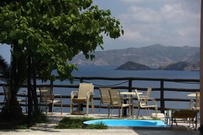 Zona de piscina con sillas y una mesa, con vista a un cuerpo de agua y montañas.