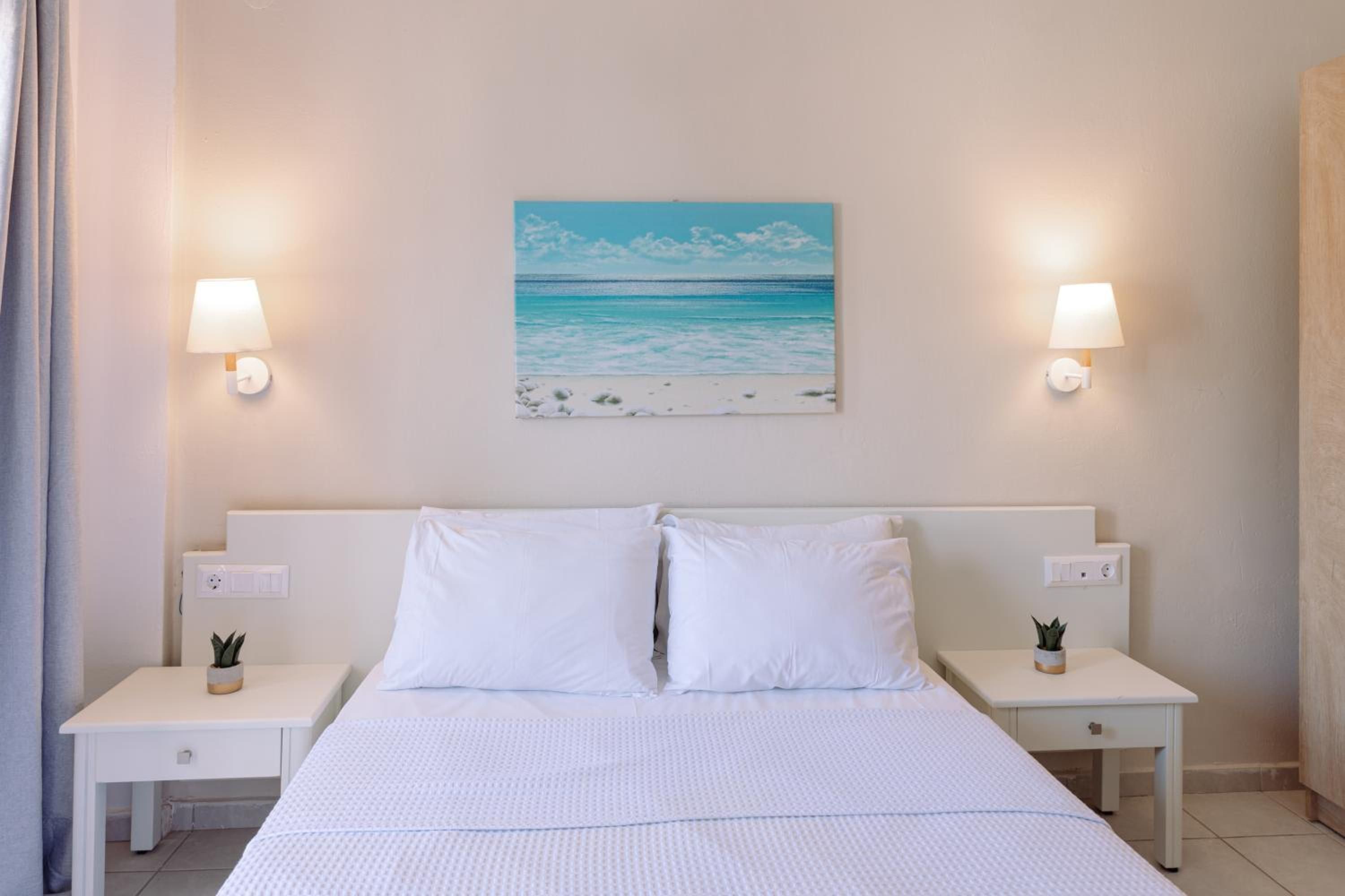 A hotel room with a bed, two bedside tables, a painting of a beach, and two wall-mounted lamps.