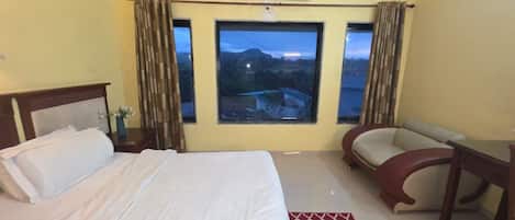 A hotel room with a bed, a desk, a chair, and a large window offering a view of the outdoors.