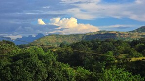 Un paisaje con un puente colgante, abundante vegetación y montañas distantes bajo un cielo parcialmente nublado.