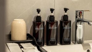 Um banheiro com pia branca, três frascos de shampoo BBLUZEN, um dosador de sabonete branco e um espelho sobre a pia.