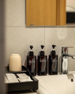 Un baño con un lavabo blanco, tres botellas de champú BBLUZEN, un dispensador de jabón blanco y un espejo sobre el lavabo.