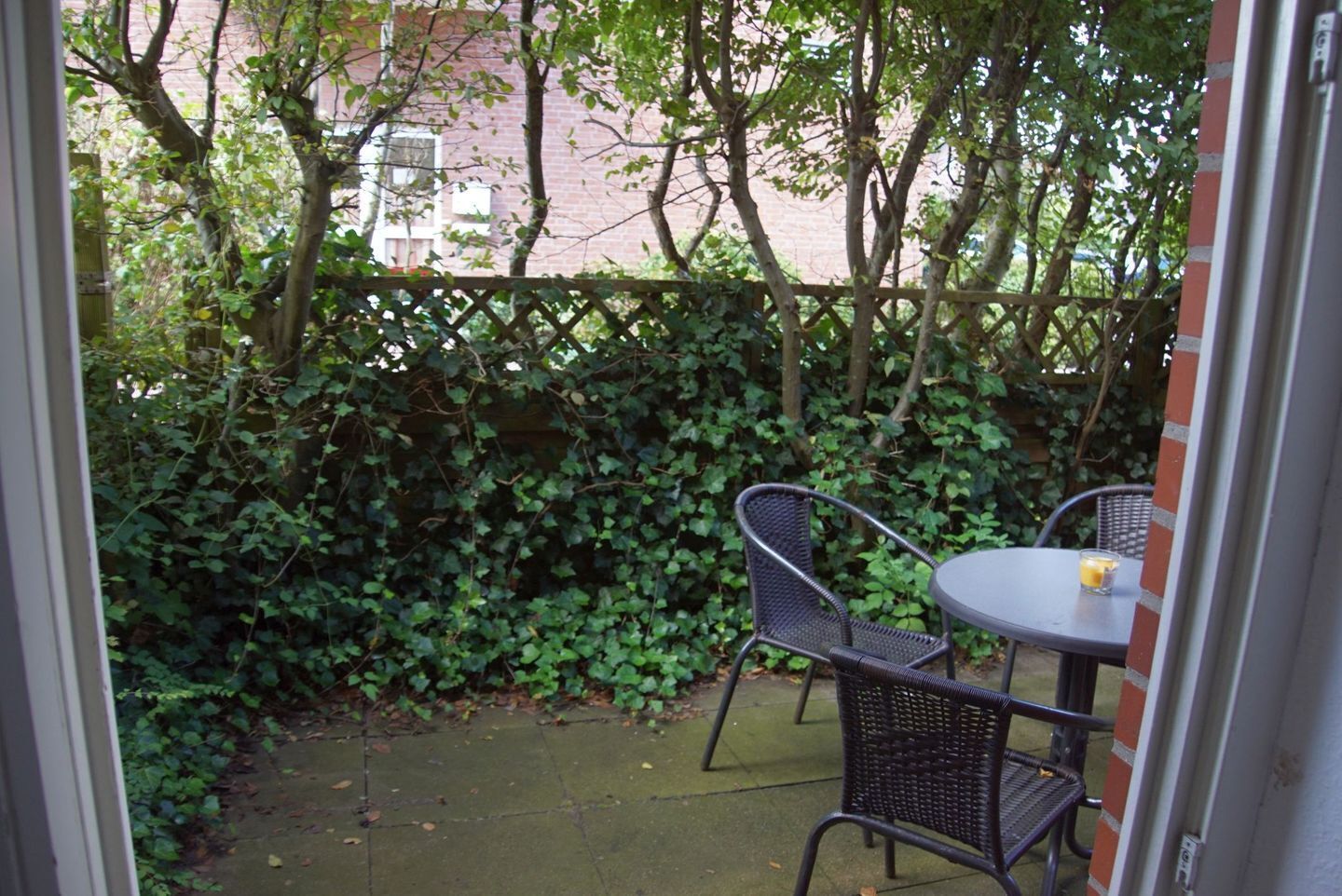 An outdoor patio with a table and two chairs, surrounded by a hedge and trees.