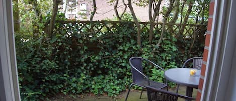 An outdoor patio with a table and two chairs, surrounded by a hedge and trees.