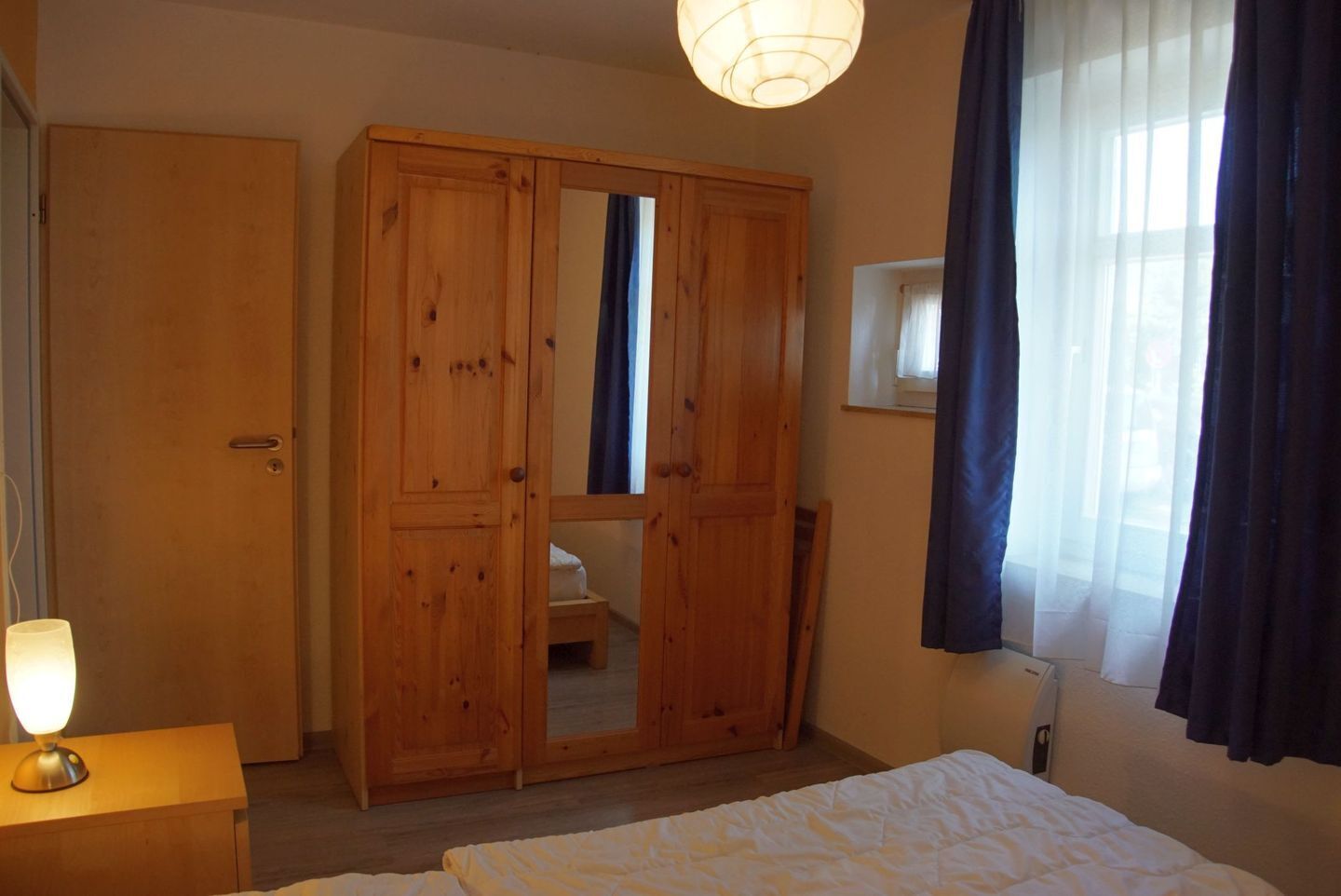Ein Schlafzimmer mit Holzschrank, Nachttisch mit Lampe, Bett mit weißer Bettwäsche und Fenster mit Vorhängen.