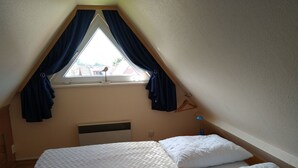 Ett sovrum med ett sluttande tak, ett fönster med gardiner, en säng med vita sängkläder och en väggmonterad värmare.