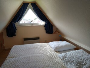 Ein Schlafzimmer mit schrägen Wänden, einem fenster mit Vorhängen, einem Bett mit weißer Bettwäsche und einem Wandheizkörper.