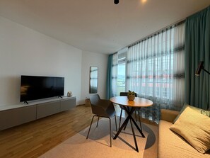A modern living room with a flat-screen TV, a dining table with chairs, a sofa, and a large window with curtains. - Romantic lakeside cabin with a balcony and indoor communal pool in Uhldingen-Mühlhof (Uhldingen-Mühlhofen)