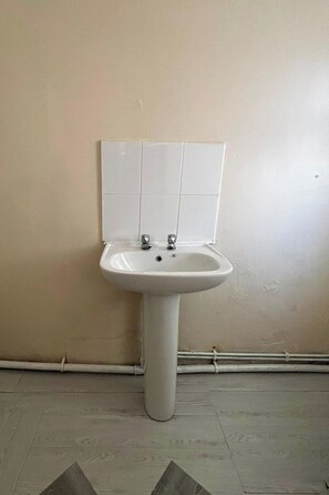 A white pedestal sink with two faucets, mounted on a beige wall above a tiled floor.