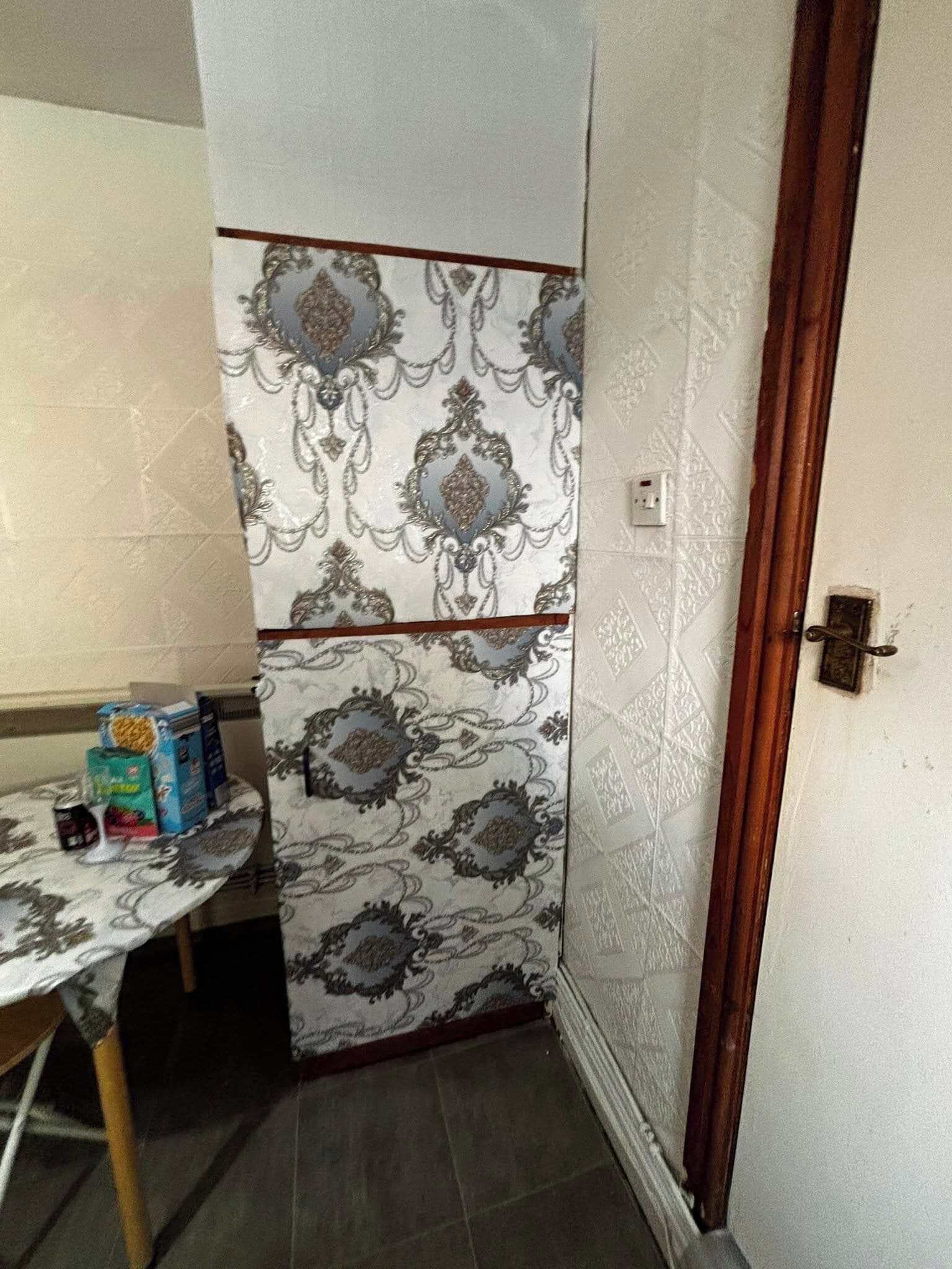 Una habitación de esquina con papel tapiz estampado, una puerta de madera y una mesa con objetos encima.
