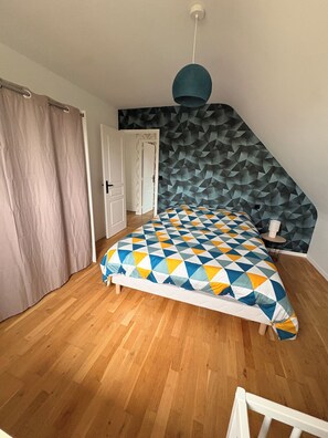Ein Schlafzimmer mit schrägen Wänden, einer Bettdecke mit geometrischem Muster, Holzboden und einer Tür zu einem anderen Raum.