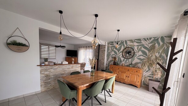 A dining area with a wooden table and chairs, a kitchen in the background, and a wall with a leaf-pattern wallpaper. - Charming house near the ocean and the village (Hillion)