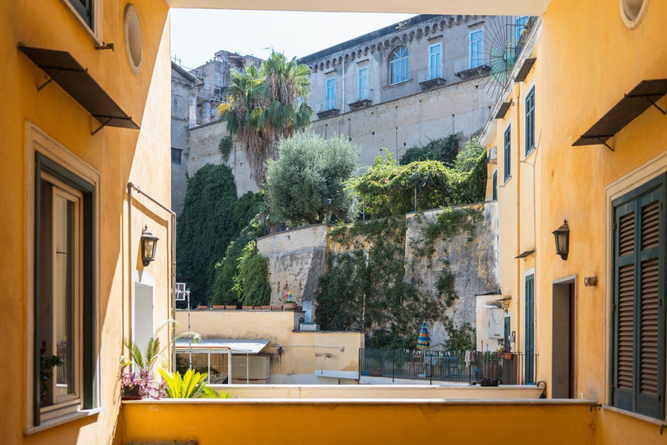 A narrow courtyard with a yellow building, a staircase, and a view of a historic stone wall with vegetation.
