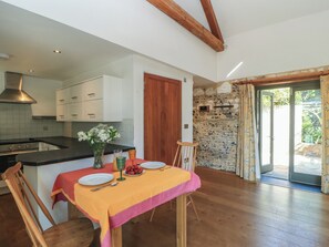 A dining area with a table set for two, a kitchen with white cabinets, and a stone wall. - Frome Gazey Barn (Dorchester)