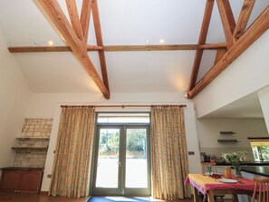 A room with a high ceiling supported by wooden beams, a sliding glass door leading outside, a dining area with a table set for two, and a kitchen area visible in the background. - Frome Gazey Barn (Dorchester)