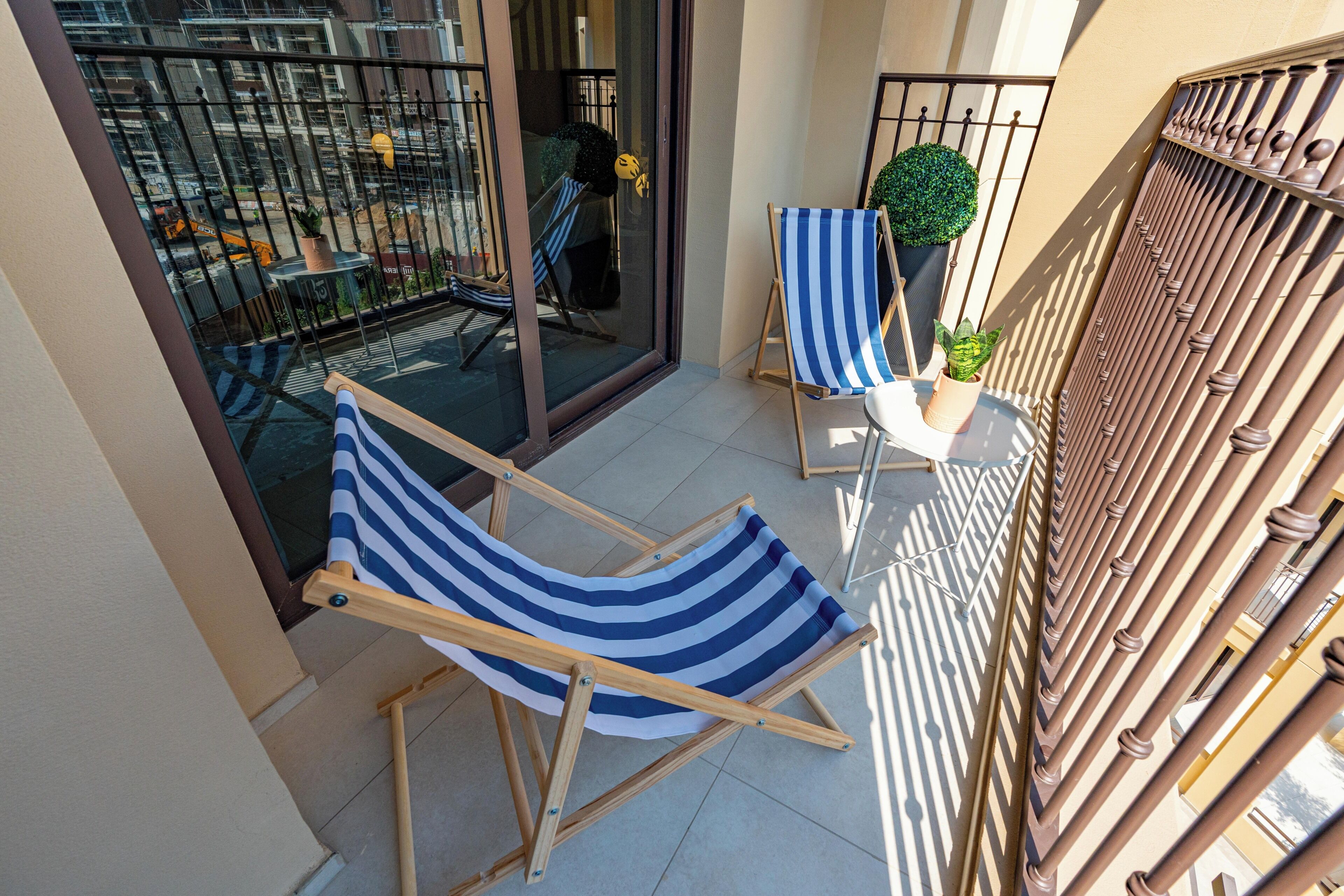 A balcony with two striped lounge chairs, a small table with a potted plant, and a view of a construction site.