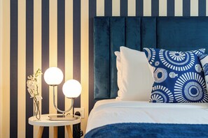 Une chambre d’hôtel avec un mur à rayures, un miroir rond, des lampes de chevet, un lit avec une tête de lit bleue et des coussins à motifs.