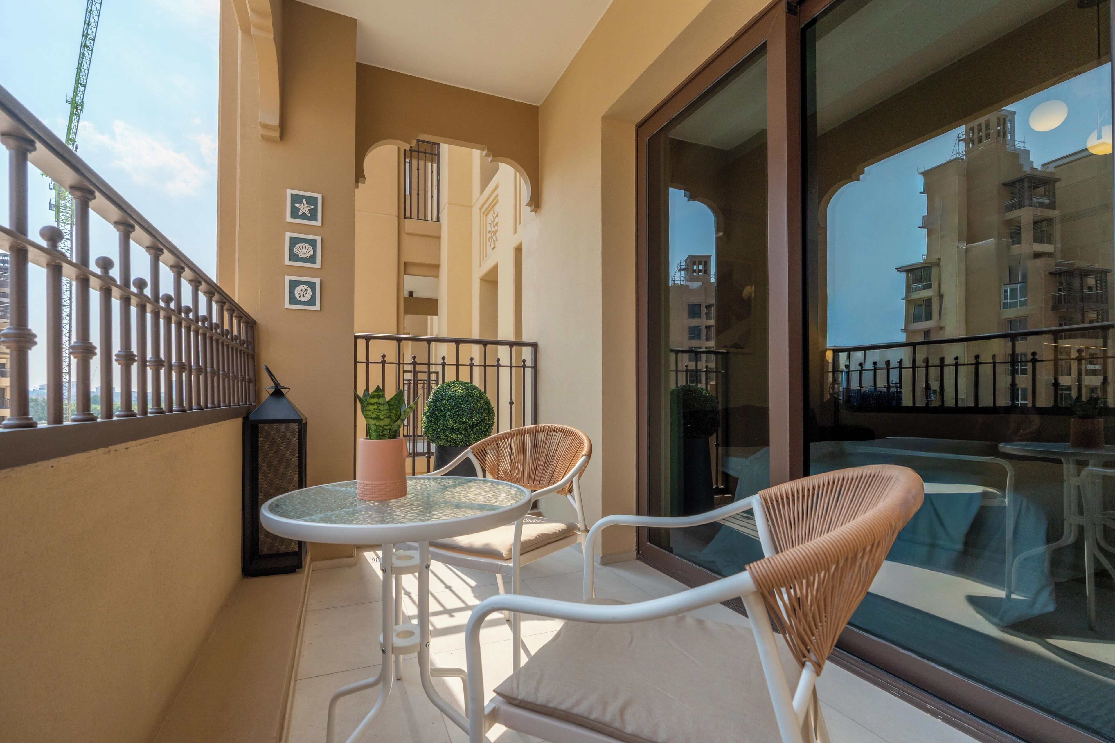 A balcony with a glass door, a round table, two wicker chairs, a potted plant, and a decorative wall with framed pictures.