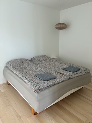 Ein Bett mit gemustertem Überwurf und zwei Kissen, Holzboden und weiße Wand mit Deckenleuchte.
