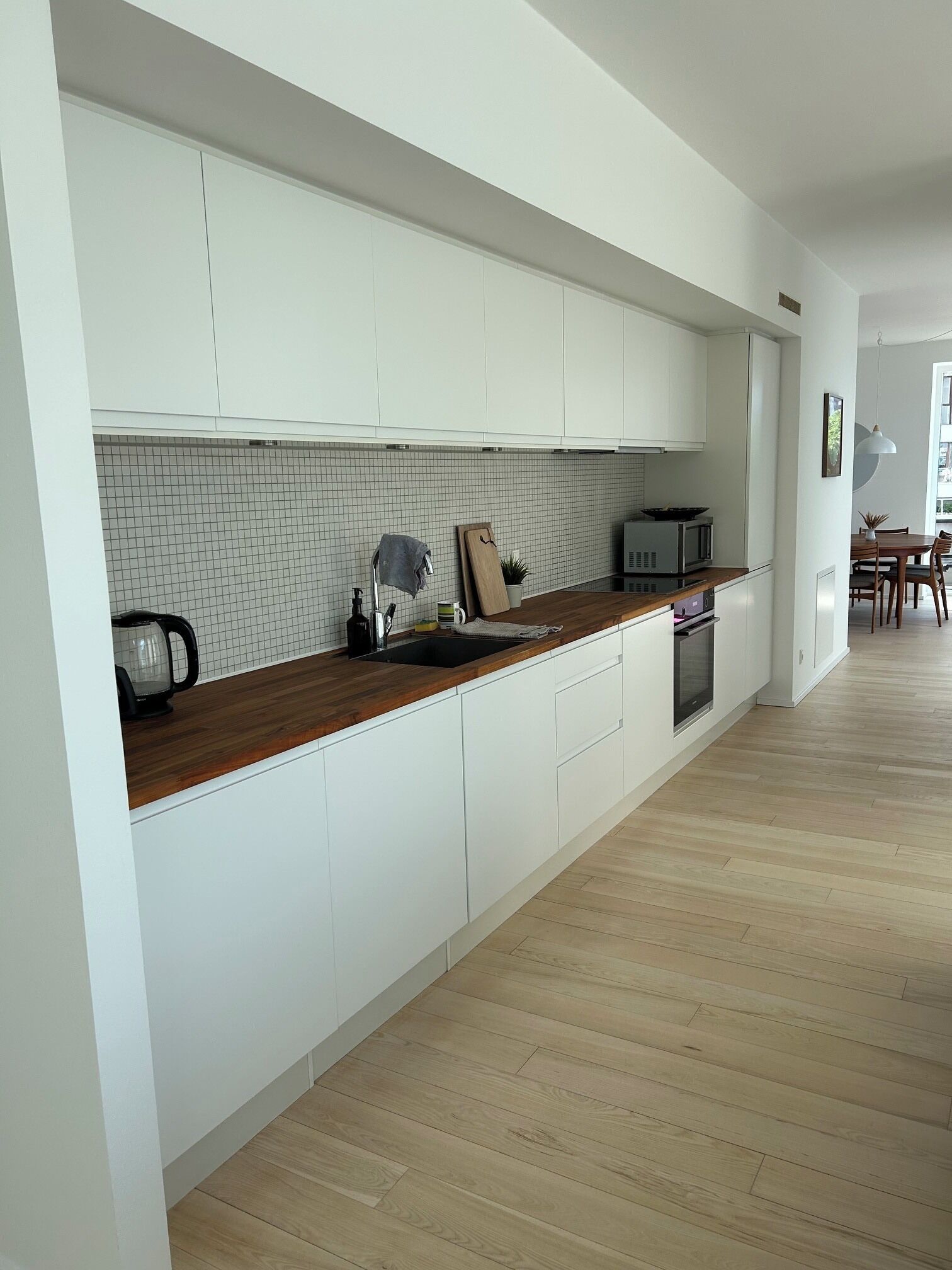 Una cocina moderna con armarios blancos, encimera de madera, una tetera, un microondas y un comedor con sillas y una mesa.