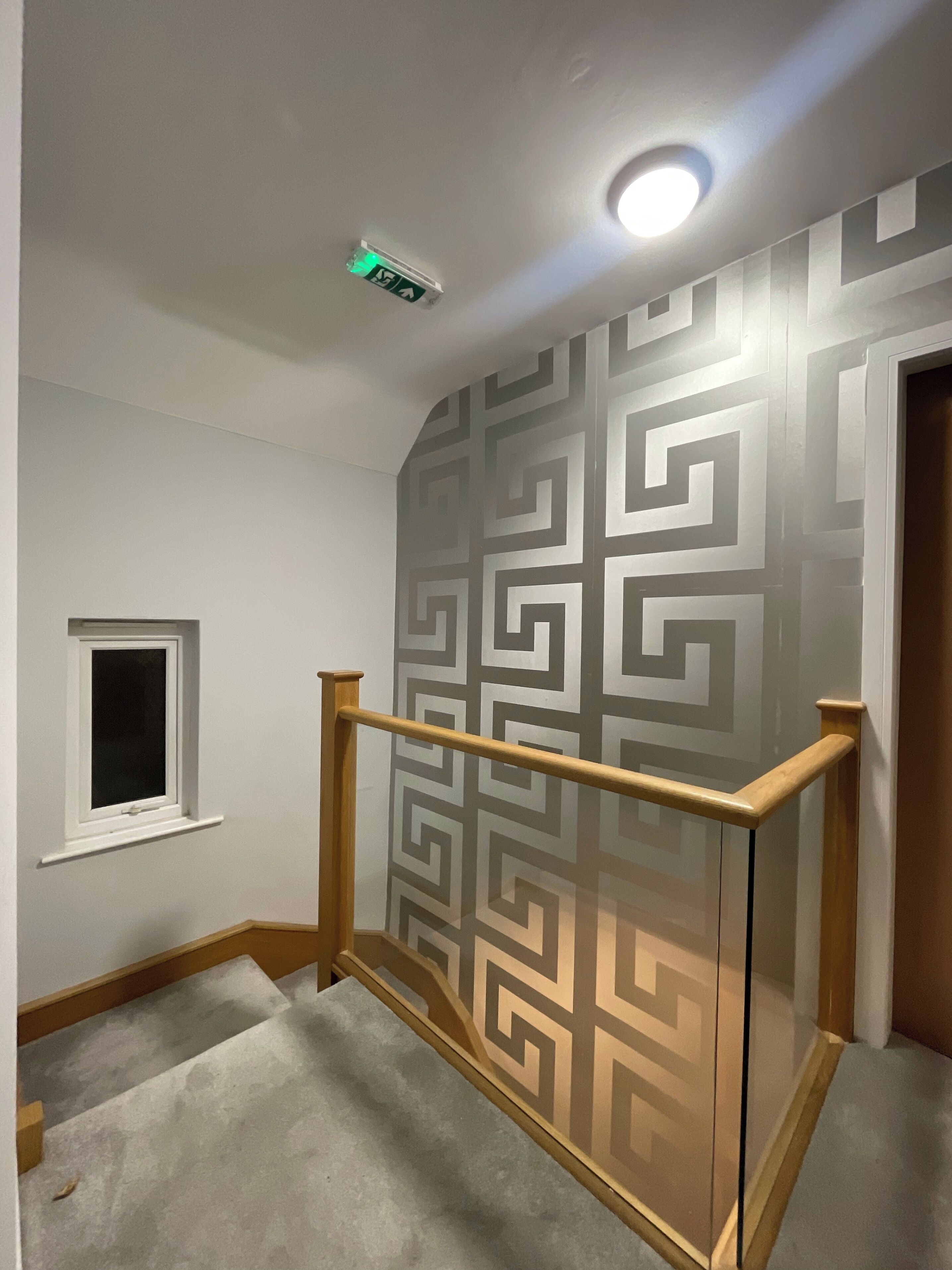 Een trap met een wand met een geometrisch patroon, een klein raam en een plafondlamp.