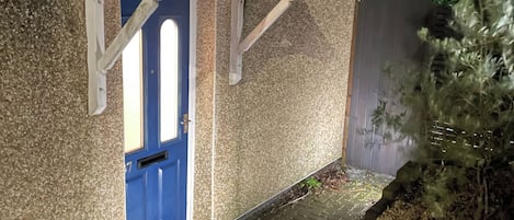 Una casa con puerta azul, un pequeño porche y un jardín con plantas y cerca de madera.
