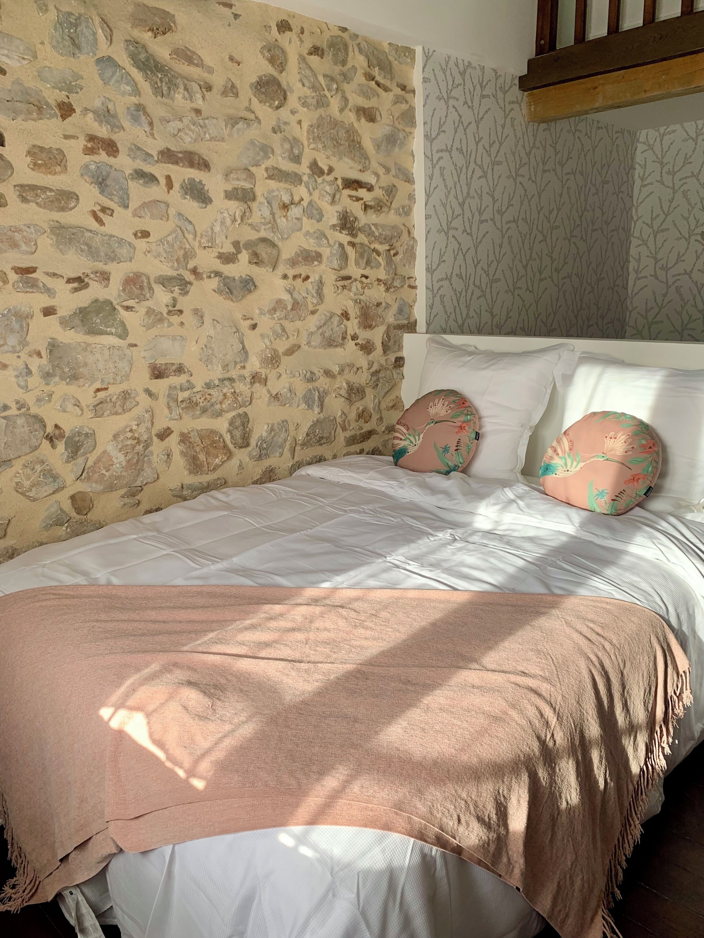 Una cama con pared de piedra de fondo, ropa de cama blanca y dos almohadas florales.