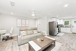 Ein modernes Wohnzimmer mit Sofa, Couchtisch, Esstisch und Küchenzeile mit Kühlschrank und Schränken.