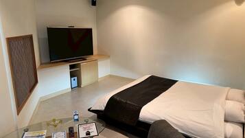 A hotel room with a bed, a television, a glass table with items, and a wall-mounted TV.