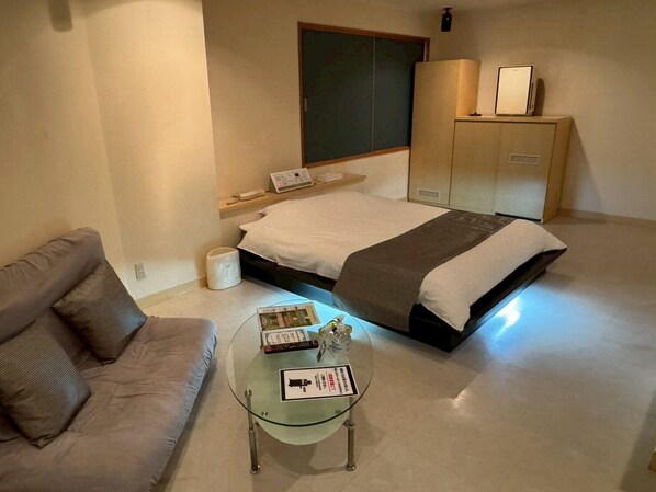 Habitación de hotel con cama, sofá, mesa de centro de vidrio, armario y mesita de noche con lámpara.