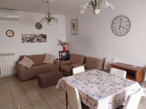 A living room with a sofa set, a dining table with a floral tablecloth, a wall clock, and a chandelier. - 
Feel right at home and relax just steps from the sea. (Pescara)