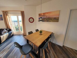 A modern living room with a wooden dining table, black chairs, a sofa, a large painting, and a clock on the wall. - Op'n Rugenbarg Vacation Rental Ebbe H60 (Cuxhaven)