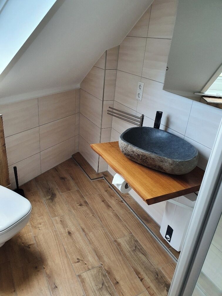 A small bathroom with a wooden floor, a white toilet, a wooden shelf with a stone basin, and a skylight.