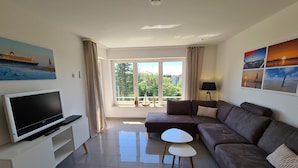 A modern living room with a flat-screen TV, a sofa, a small table, and a balcony with a view of greenery. - Op'n Rugenbarg Vacation Rental Elbe H64 (Cuxhaven)