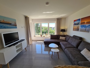 Ein modernes Wohnzimmer mit einem Flachbildfernseher, einer Couch, einem kleinen Tisch und einem Balkon mit Blick auf Grünflächen.