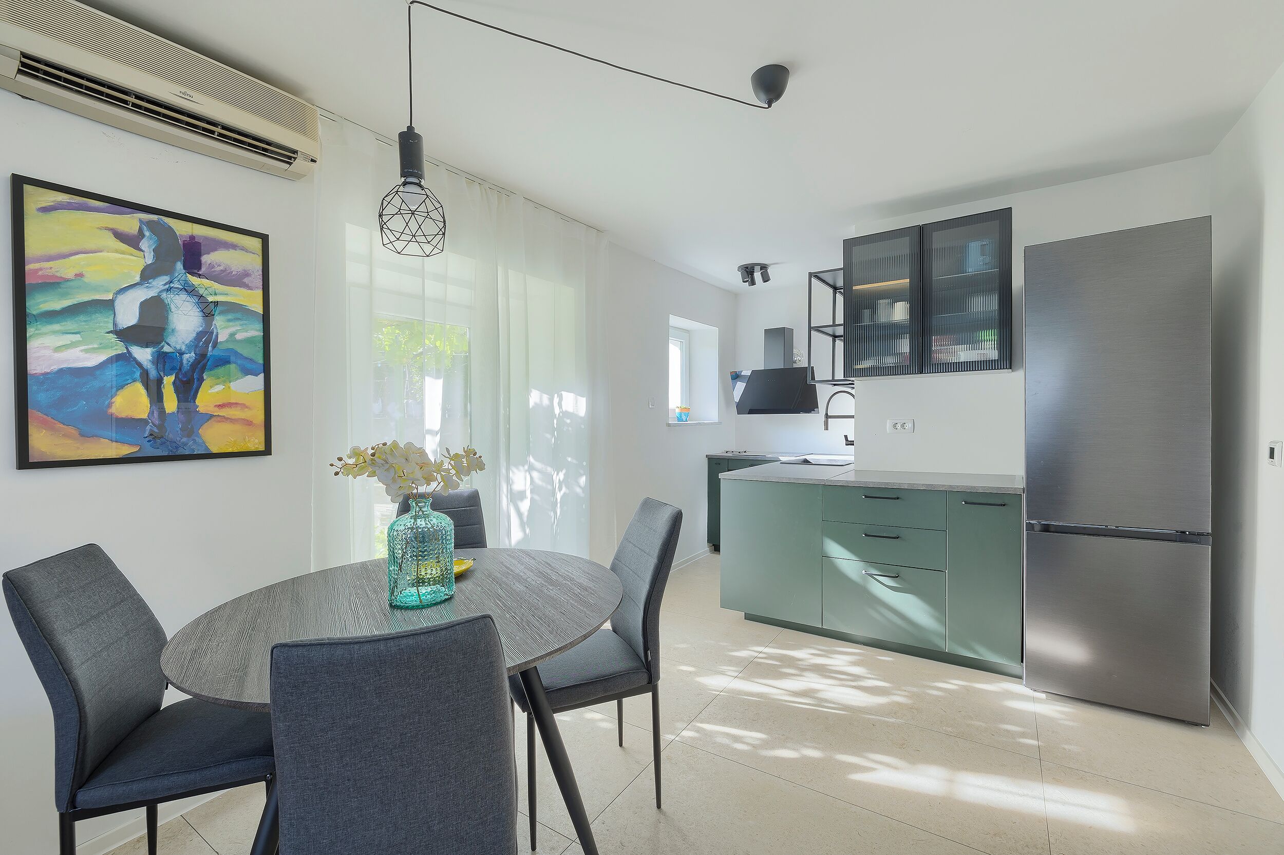 A modern kitchen with a stainless steel refrigerator, a dining area with a round table and chairs, and a colorful abstract painting on the wall.