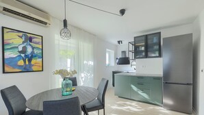 A modern kitchen with a stainless steel refrigerator, a dining area with a round table and chairs, and a colorful abstract painting on the wall. - Fortica Garden Apartment – Pula Arena, Free Parking (Pula)