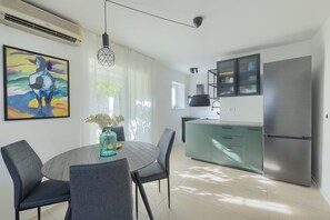 A modern kitchen with a stainless steel refrigerator, a dining area with a round table and chairs, and a colorful abstract painting on the wall. - Fortica Garden Apartment – Pula Arena, Free Parking (Pula)