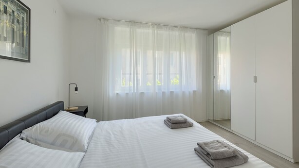 A hotel room with a bed, a nightstand, a lamp, a window with sheer curtains, and a door leading to a closet. - Fortica Garden Apartment – Pula Arena, Free Parking (Pula)