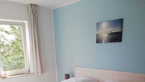 A bedroom with a bed, a nightstand, a lamp, a window with curtains, and a wall-mounted picture. - Op'n Rugenbarg Vacation Rental, Wadden Sea, H62 (Cuxhaven)
