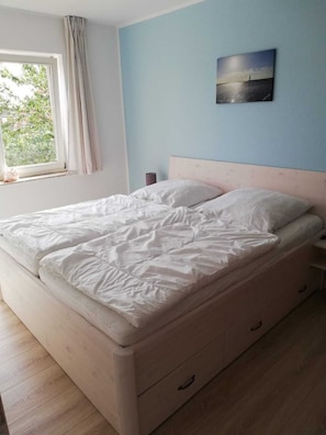 Ein Schlafzimmer mit einem Bett, einem Nachttisch, einer Lampe, einem Fenster mit Vorhängen und einem an der Wand hängenden Bild.