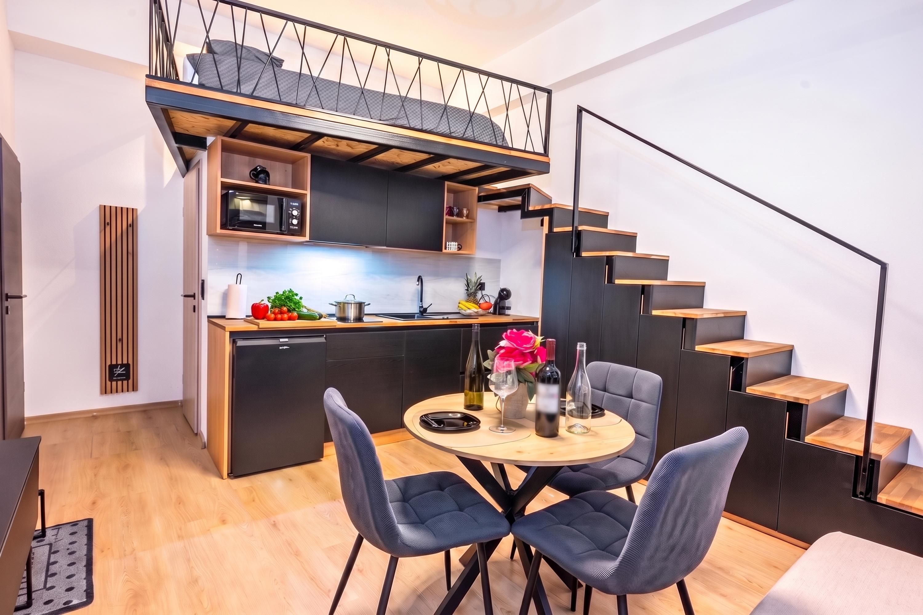 A compact kitchen with a microwave, sink, and wooden cabinets. There is a dining area with a round table and two chairs. A staircase with wooden steps leads to an upper level.