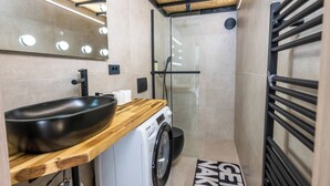 A compact bathroom with a wooden vanity, a black sink, a washing machine, and a glass shower enclosure. - Alter 26 Urban Apartment With Free Parking (Koper)
