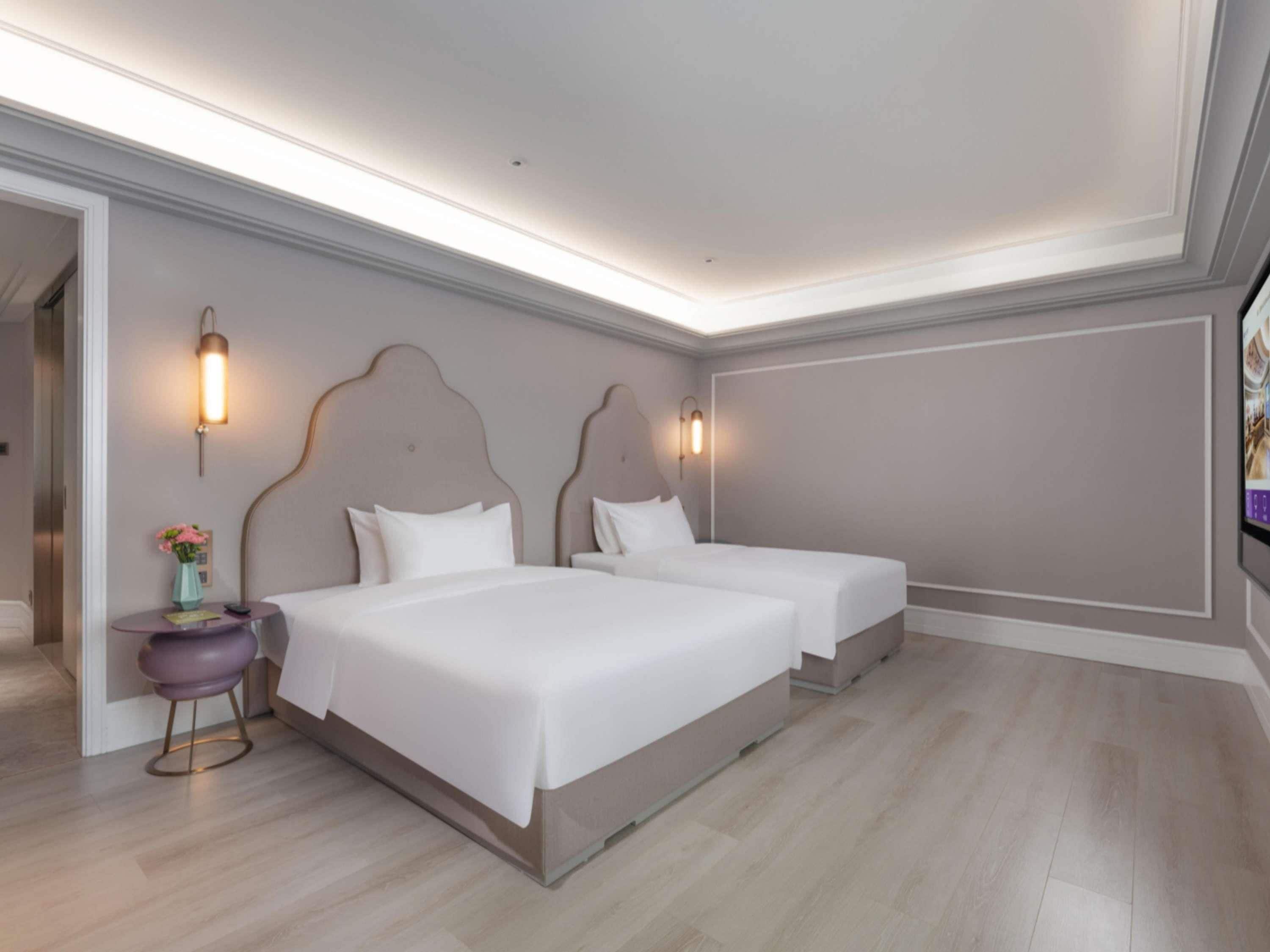 A modern hotel room with two beds, a small round table with a vase, and a TV on the wall.