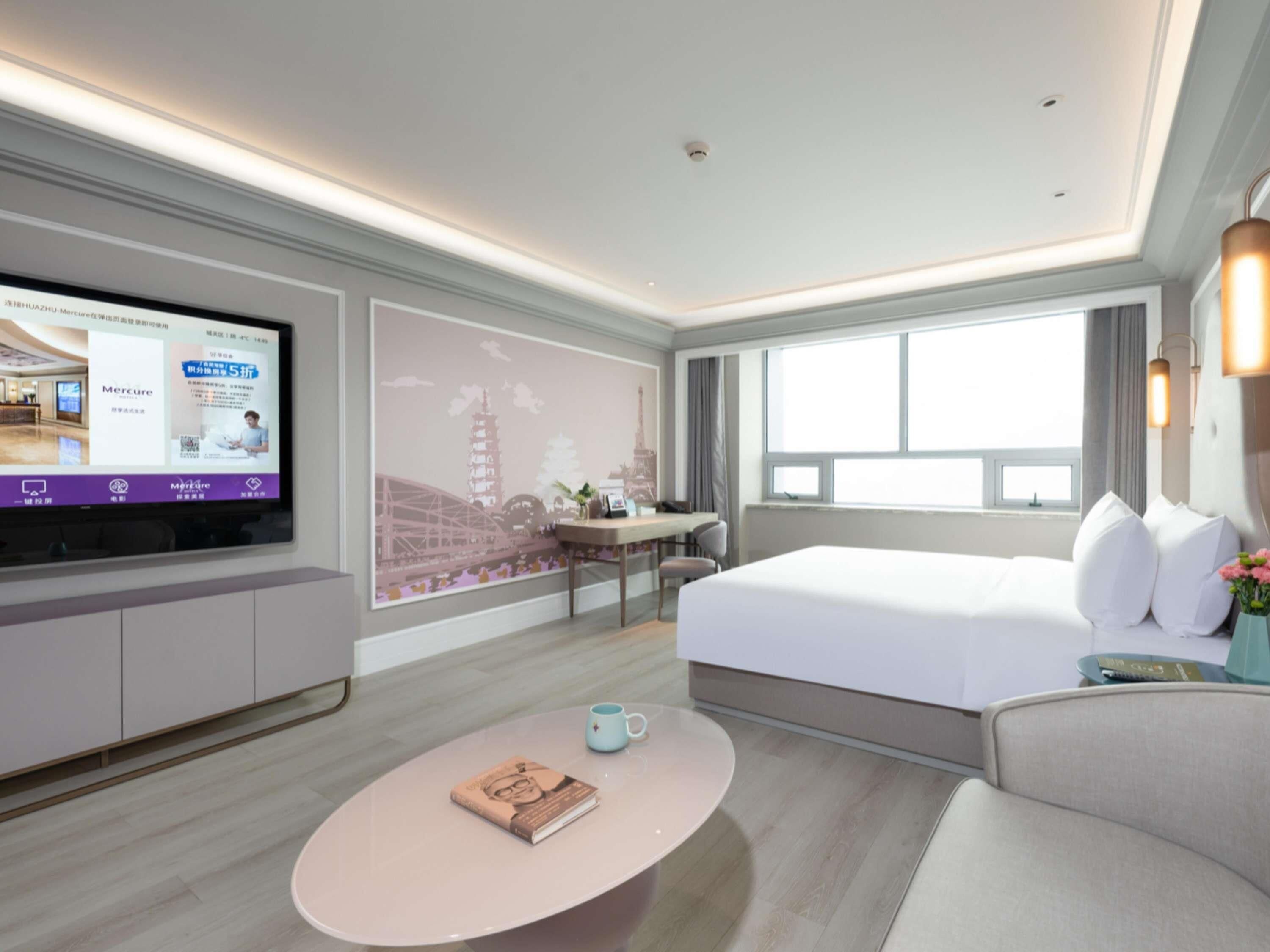 A modern hotel room with a large bed, a flat-screen TV mounted on the wall, a round coffee table, and a desk with a chair.