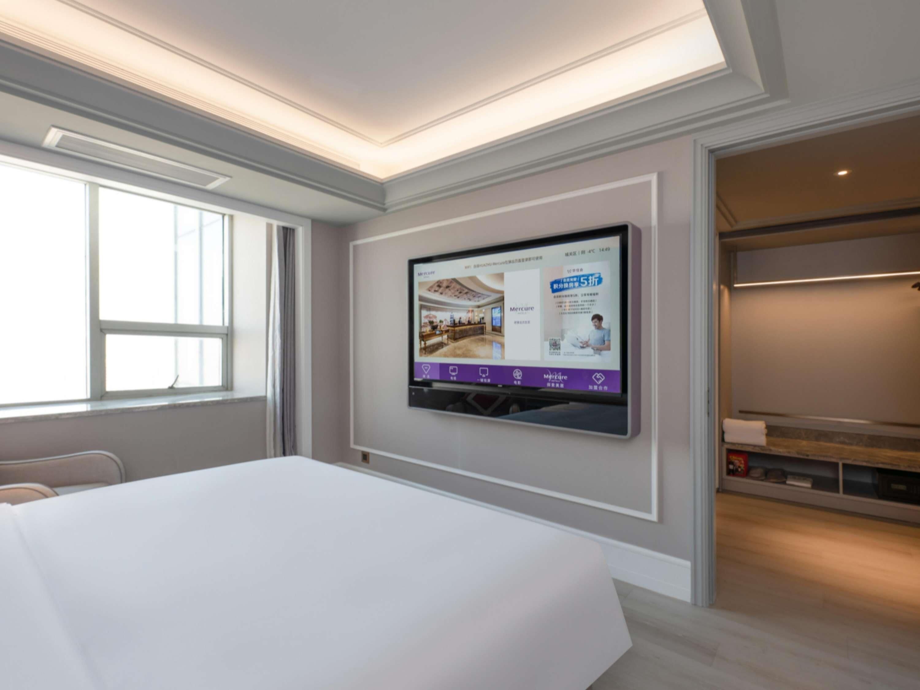 A hotel room with a large bed, a flat-screen TV mounted on the wall, and a window providing natural light.
