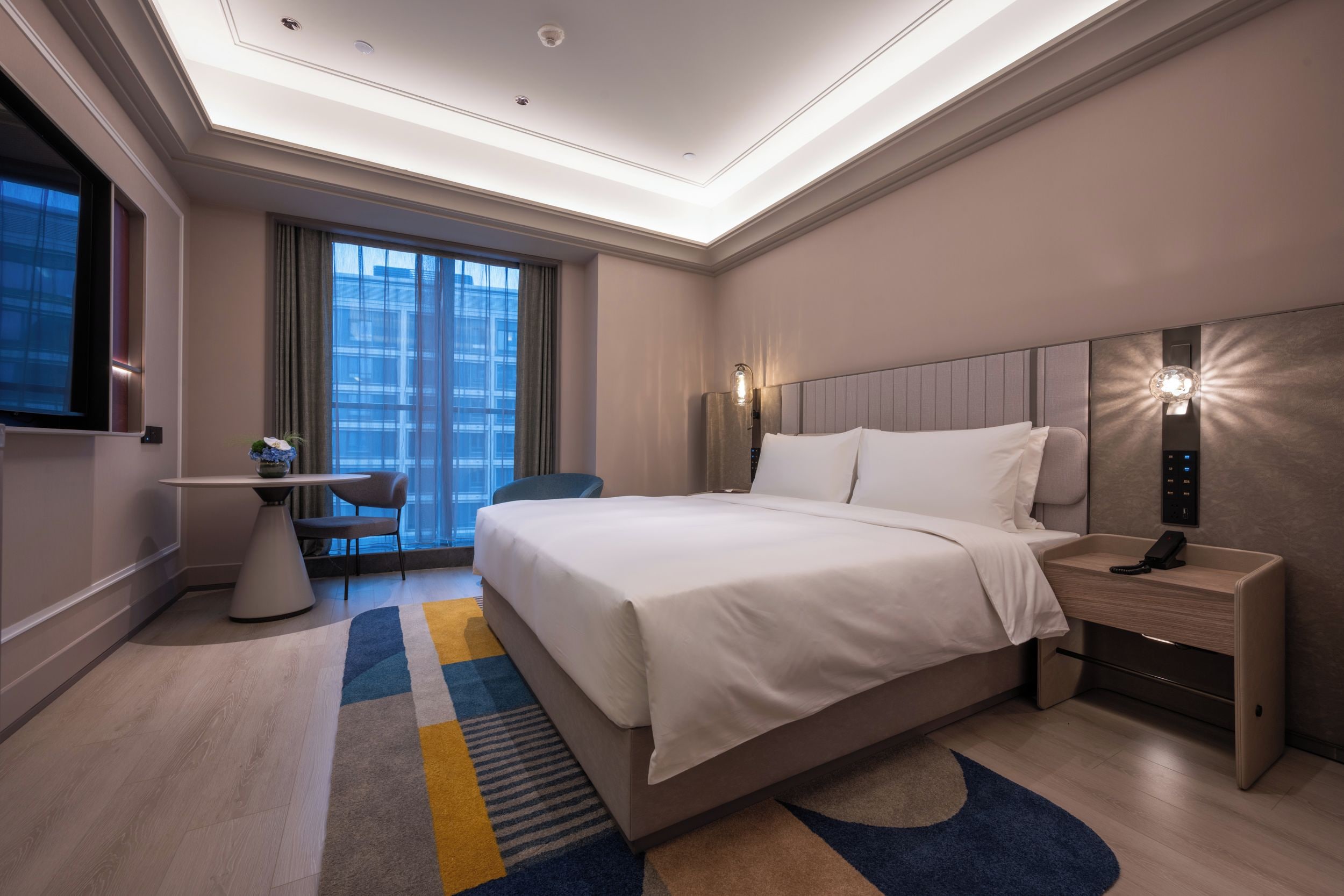 A modern hotel room with a large bed, a desk, a chair, and a view of the cityscape through the window.