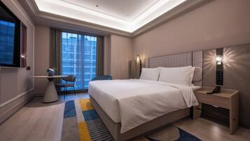 A modern hotel room with a large bed, a desk, a chair, and a view of the cityscape through the window.